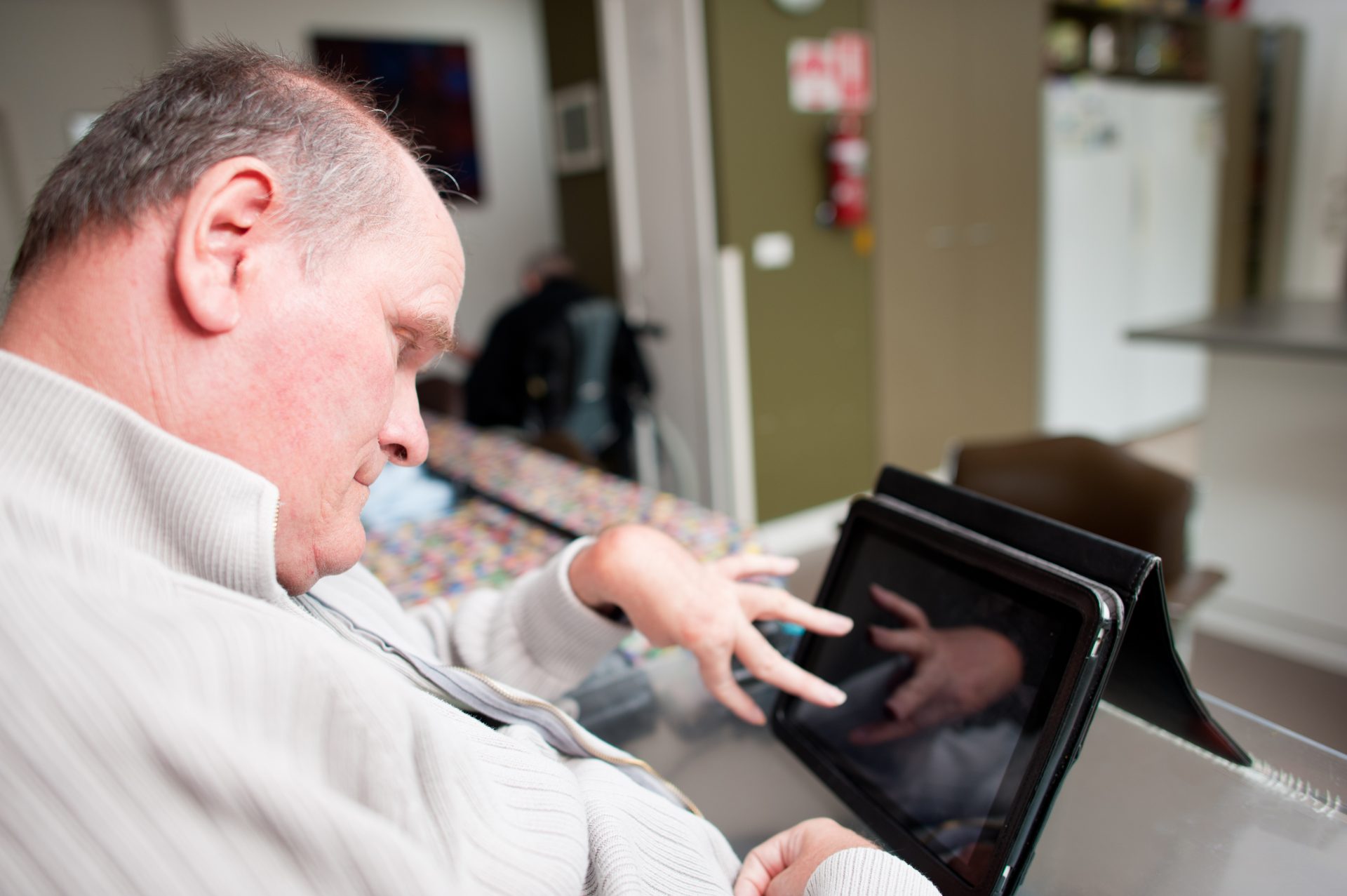 Mature aged man with a disability operating touchscreen computer.  The reflection of his hand can be seen on the screen.  He is seated in his living room.