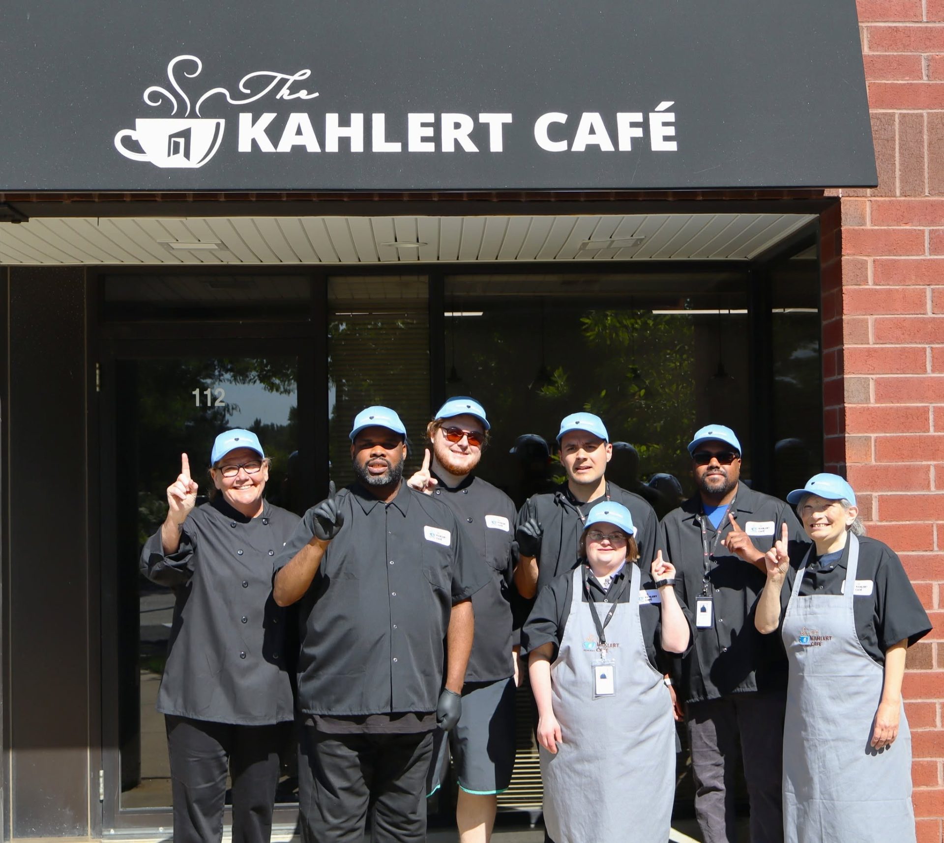 Group of 7 people wearing chef aprons and hats in front of cafe