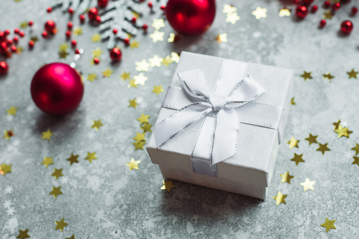 Silver gift with bow on grey snowy background with red Christmas balls and gold confetti stars.